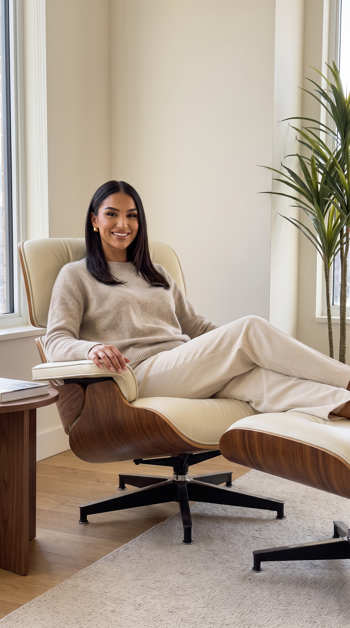 A woman sits comfortably in a modern mid-century style lounge chair with a matching ottoman in a bright, neutral-toned room. 